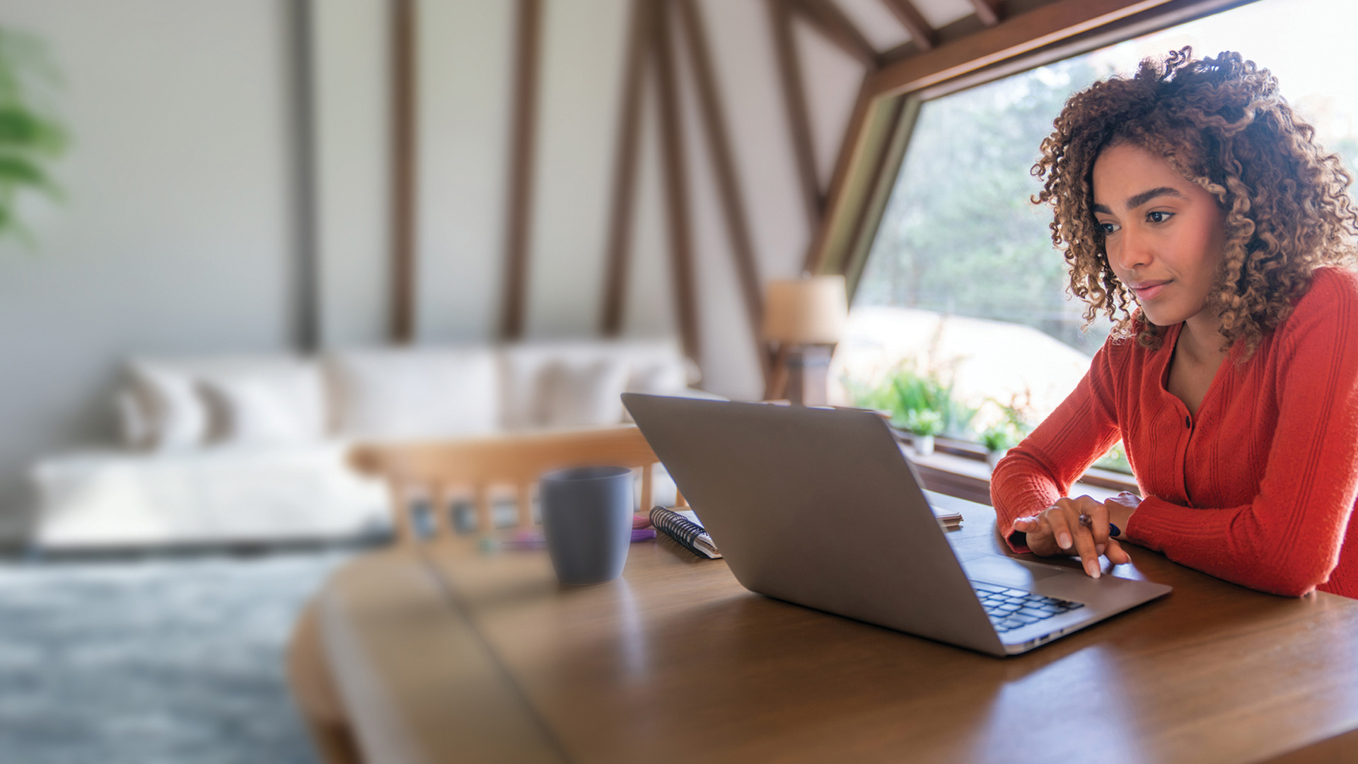 A woman with curly hair sits at a wooden table in a bright room, working on a laptop. A coffee mug is placed beside the computer, and large windows reveal greenery outside.