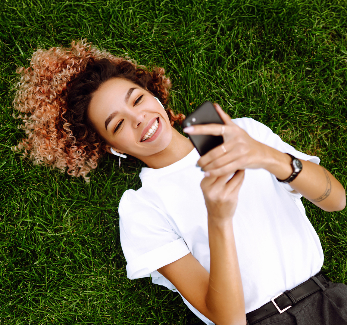 Close-up of a person lying on green grass, holding a smartphone and wearing earbuds, with curly hair spread out on the ground and dressed in a white shirt.